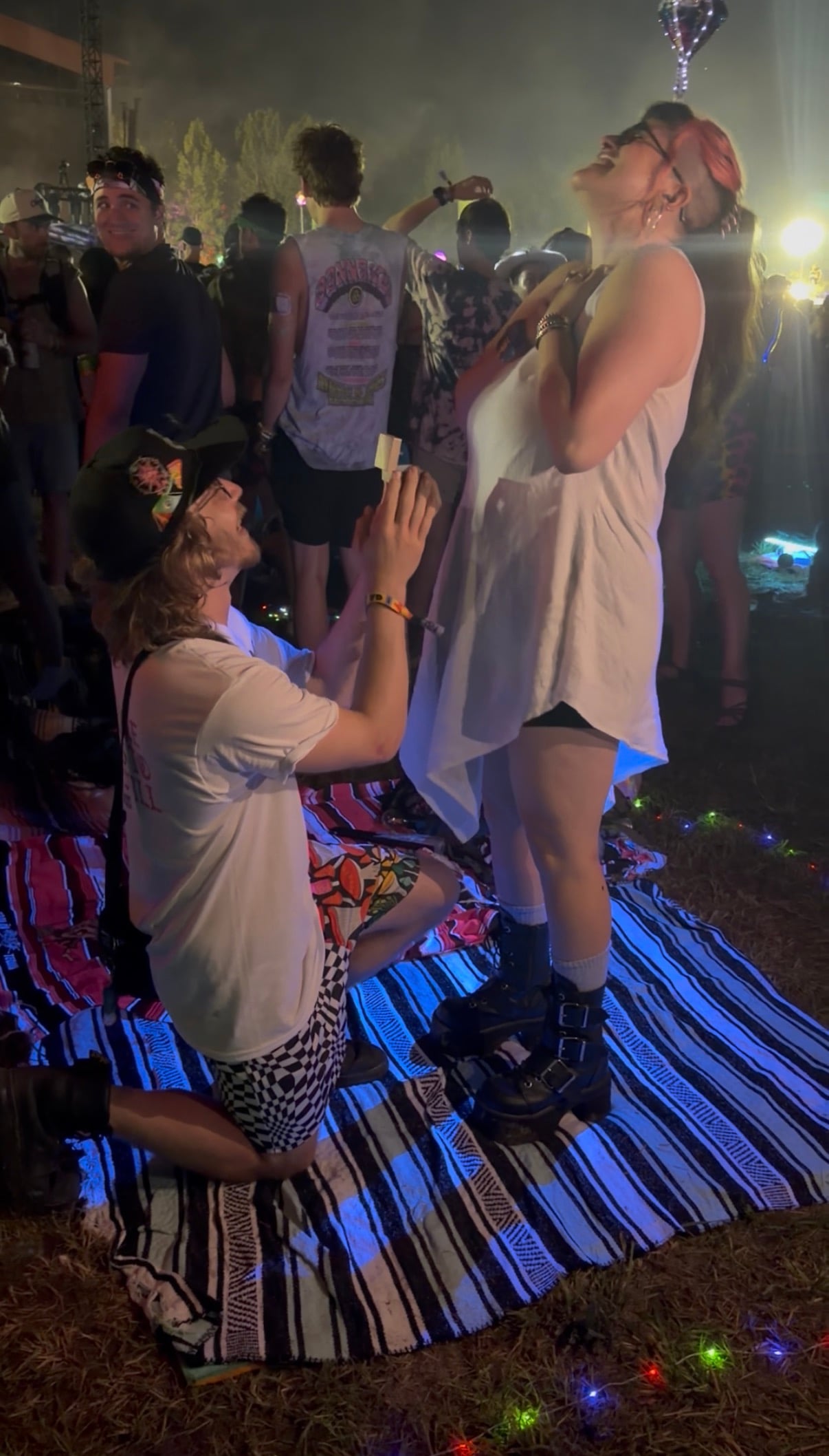 A proposal on one knee at Bonnaroo, festival lights and smoke behind.